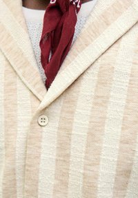 Blazer rayé beige et blanc en tissu texturé, avec une fermeture à bouton unique et une écharpe rouge autour du cou.
