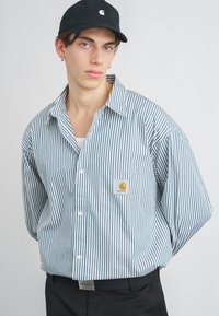 Camisa de manga larga a rayas en azul claro y blanco, con un bolsillo en el pecho que presenta un logo, combinada con una gorra negra. Ajuste casual y relajado.
