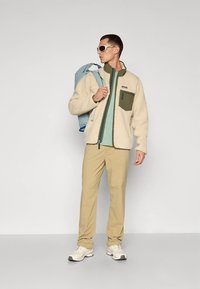 Kreemika värvi fliisjakk rohelise tasku ja lukuosaga, kombineeritud helerohelise kihiga, beežide pükste ja valgete tossudega. Komplektis on hele sinine seljakott.