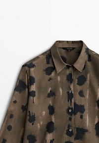 Kaulusmainen napillinen paita ruskeasta kankaasta, jossa on mustia abstrakteja pilkkuja. Ominaisuuksiin kuuluu pehmeä pinnanrakennus ja rento istuvuus.