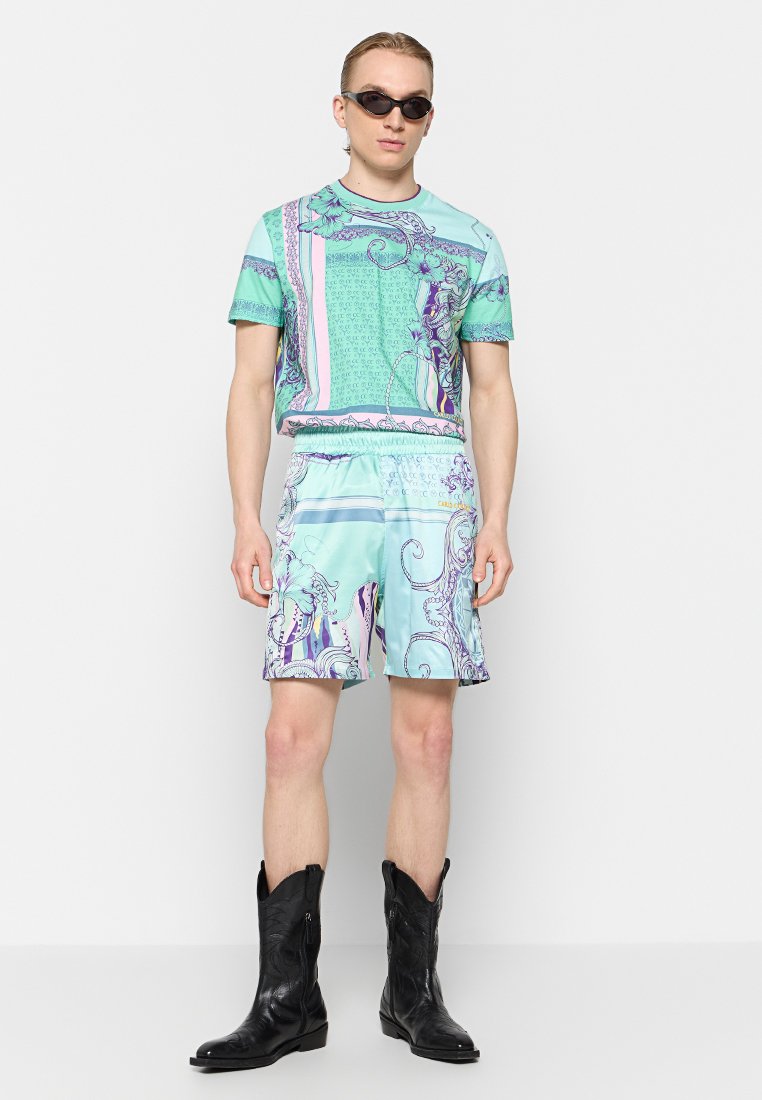 Kurzärmeliges, mintgrünes Hemd und passende Shorts mit komplexen lila und rosa Mustern. Getragen mit schwarzen Stiefeln im Cowboy-Stil und Sonnenbrille.