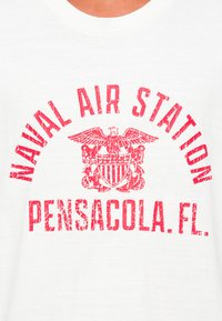 Raudonas nusidėvėjęs tekstas "Naval Air Station Pensacola, FL" su erelio emblema ant balto audinio fono.