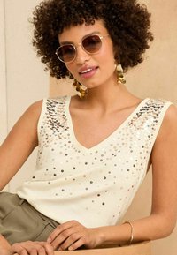 Ivory sleeveless top with a V-neck, embellished with sequins in various shades, paired with olive green trousers and gold accessories.
