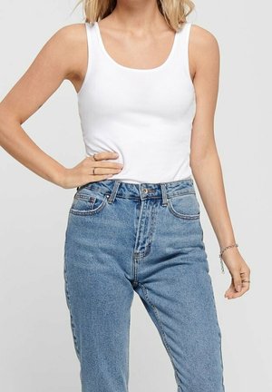 Femme portant un débardeur blanc sans manches et un jean bleu taille haute, main sur la hanche et bracelet léger au poignet.