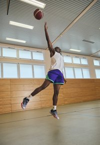 En atlet iført en hvid tanktop og lilla shorts med grønne detaljer hopper for at skyde en basketball i et gym med trægulve og klart lys.