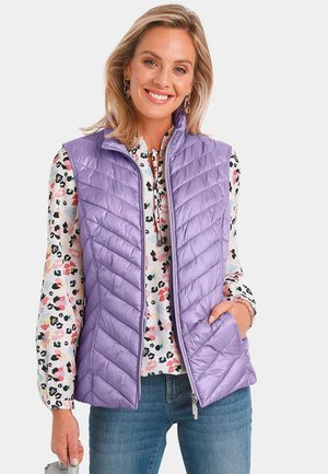 Lila gesteppte Weste mit Reißverschluss, hohem Kragen und seitlichen Taschen, über einer mehrfarbigen Bluse mit Blumenmuster getragen, kombiniert mit Jeans.