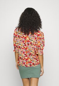 Blusa com padrão floral, com mangas curtas bufantes, em cores vibrantes como vermelho e amarelo, com um corte descontraído. Combinada com uma saia verde.