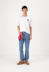 White short-sleeve t-shirt with a blue and red graphic, light blue denim jeans, red bandana tied at the waist, brown sneakers with laces.