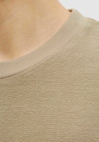 Beige striktrøje stof med en tekstureret overflade og en ribbet rund halsudskæring. Farven er ensartet i hele materialet.