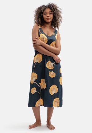 Donna con capelli ricci indossa un vestito blu scuro senza maniche con grandi motivi di foglie gialle, in piedi a piedi nudi con le braccia incrociate.