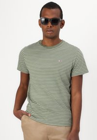 Camiseta a rayas verdes con cuello redondo, manga corta y un pequeño detalle de logo. Se lleva con gafas de sol oscuras y pantalones beige.