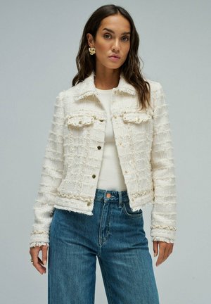 Femme portant une veste blanche texturée à boutons sur un haut blanc, associée à un jean bleu taille haute, debout devant un fond uni.