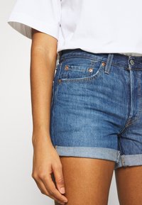Shorts en denim d'un bleu moyen avec des ourlets retroussés, présentant des accessoires en cuivre, des détails de couture et un design classique à cinq poches.