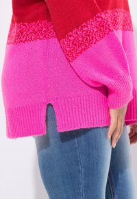 Primo piano di una persona che indossa un maglione lavorato a maglia rosa e rosso con orlo a spacco, abbinato a jeans blu, con la mano poggiata sul fianco.