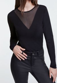 Vrouw in een zwart langemouwtop met een doorschijnende V-vormige halslijn, gecombineerd met hooggetailleerde zwarte leren broeken, hand rustend op de heup.