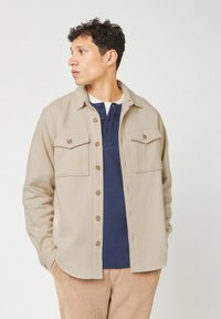 Beige button-up jas met twee borstzakken, gebogen zoom en bruine knopen. Draagt zich over een donkerblauwe henley shirt, gepresenteerd tegen een neutrale achtergrond.