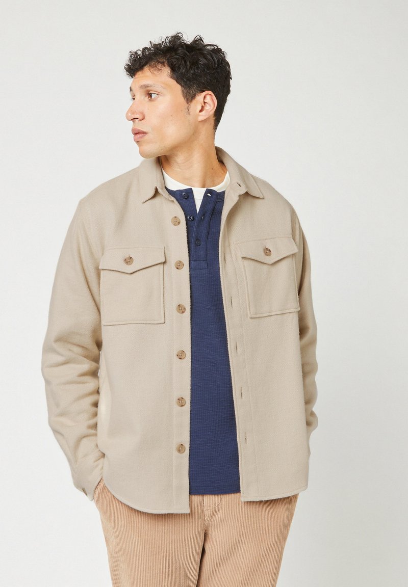 Beige button-up jas met twee borstzakken, gebogen zoom en bruine knopen. Draagt zich over een donkerblauwe henley shirt, gepresenteerd tegen een neutrale achtergrond.