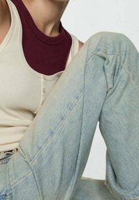 Sopivuuden beige hihaton paita, jossa on syvä viininpunainen kaula-aukko; vaaleansiniset korkeavyötäröiset farkut, joissa on hieman haalistunut pinta ja näkyvät saumat.