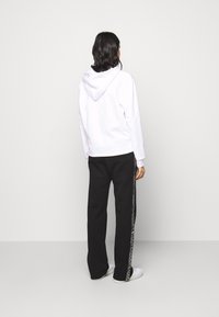 Donna in piedi, di spalle, con un felpa bianca, pantaloni neri con strisce laterali e sneakers bianche, su uno sfondo semplice.