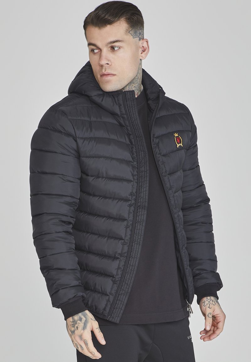 Veste rembourrée noire avec un design matelassé, capuche amovible, accent brodé sur la poitrine et poignets côtelés. Dispose d'une fermeture éclair à l'avant.