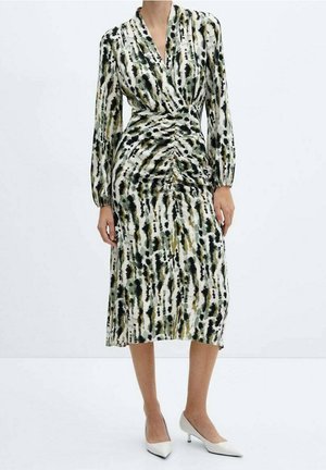 Femme portant une robe midi à manches longues, froncée, avec un imprimé abstrait noir, vert et beige, chaussée de escarpins blancs à bout pointu et talons kitten.