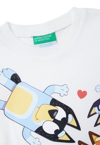 Biała bawełniana koszulka z grafiką kreskówkowej postaci z niebieskimi i czarnymi akcentami. Zawiera zieloną metkę i czerwone serce jako detal.