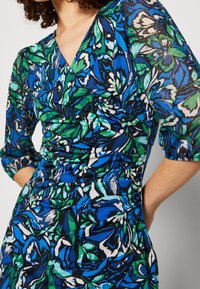 Vestido cruzado floral con patrones en verde y azul con acentos negros. Confeccionado con tela ligera, tiene mangas de tres cuartos y cintura entallada.