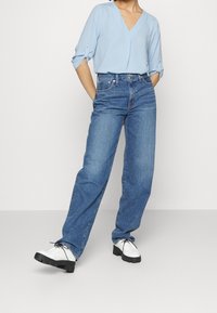 Persona che indossa una blusa azzurra a V con maniche arrotolate, jeans a vita alta e a gamba larga blu, e scarpe bianche con lacci e suola nera.