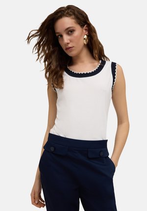 Haut sans manches blanc avec col et emmanchures en forme de vagues bleu marine, associé à un pantalon taillé bleu marine avec poches avant et accents de boutons.