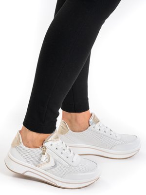 Persona che indossa sneakers bianche traforate con accenti beige e argento, leggings neri, in piedi su una superficie bianca.