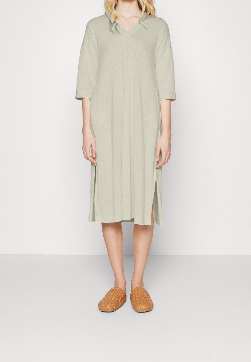 Femme portant une robe ample vert clair longueur genoux avec manches trois-quarts et chaussures tissées beiges, debout devant un fond uni.