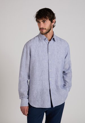 Mann trägt hellblaues, fein gestreiftes Hemd mit langen Ärmeln und Kragen, kombiniert mit dunkelblauer Hose, steht vor einfachem Hintergrund.