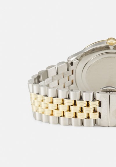 Silver and gold metal watch band with layered woven design. Features polished links and a clasp with embossed logo detail. Round watch back visible.