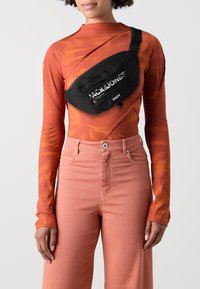 Black crossbody bag with a textured finish and logo print, worn over an orange patterned long-sleeve top and paired with pink high-waisted jeans.