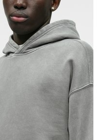 Η φωτεινή γκρι κουκούλα είναι κατασκευασμένη από μαλακό ύφασμα, διαθέτει κορδόνι για τη κουκούλα, ριμπ μανσέτες και χαλαρή εφαρμογή με ορατές ραφές.