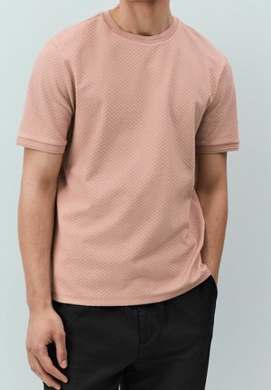 Mężczyzna ubrany w teksturowaną jasnoróżową koszulę z krótkim rękawem, z prążkowanym kołnierzem i mankietami, w połączeniu z czarnymi spodniami, stojący na jednolitym tle.