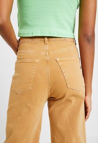 Tan, high-waisted jeans met een relaxed fit. Heeft twee achterzakken met een gestikt design. Materiaal lijkt zacht en denimachtig.