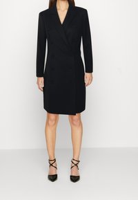 Svart dubbelknäppt kappa med slagkrage, långa ärmar och knapprad, kombinerad med svarta remsandaler med spetsig tå.