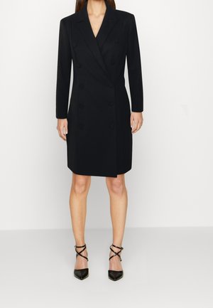 Manteau noir croisé avec un col cranté, des manches longues et des détails de boutons, associé à des escarpins noirs à bout pointu avec des brides.