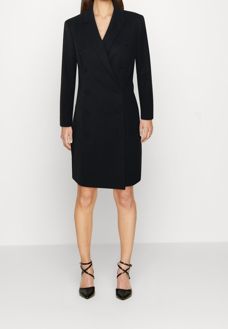 Svart dubbelknäppt kappa med slagkrage, långa ärmar och knapprad, kombinerad med svarta remsandaler med spetsig tå.