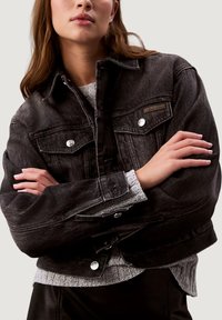 Černá džínová bunda se stříbrnými zapínacími knoflíky, dvěma náprsními kapsami a krátkým střihem. Nositelná přes šedý texturovaný svetr.