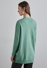 Pull en maille vert clair, coupe décontractée, avec des poignets et un ourlet côtelés. Texture douce, encolure ronde, et design du dos allongé.