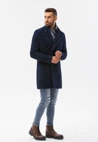 Marineblå uldfrakke med dobbeltradet design, prydet med store knapper. Kombineret med slidte lyse jeans og brune snørebåndsstøvler.