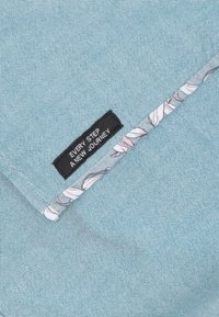 Blauwe textuurstof met een zwart label waarop staat "IEDERE STAP EEN NIEUWE REIS." Bloemmotieven accentueren een contrasterende bies aan de rand.
