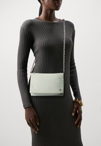 Borsa a tracolla verde chiaro realizzata in materiale liscio, con tracolla a catena e un sottile dettaglio del logo sulla parte anteriore. Abbinata a un vestito in maglia a costine.