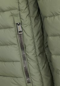 Padded grön jacka med en texturerad yta, som har en svart dragkedja med en dragflik på sidan och sydda detaljer för struktur.
