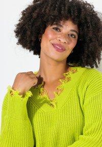 Pull en maille de couleur vert lime vif avec un décolleté en V. Il présente des bords effilochés, un tissu texturé et des manches longues à coupe ample.