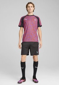 Puma sporta krekls violetā un melnā krāsā ar viļņotu rakstu, kopā ar melnām sporta biksēm, melnām zeķēm līdz ceļiem un rakstainiem futbola apaviem.