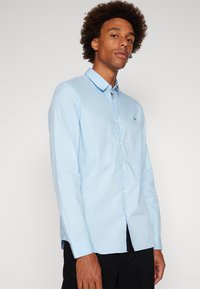 Helles blaues Langarmhemd mit Button-Down-Kragen und kleinem grünen Logo auf der Brust. Der Stoff wirkt glatt und leicht strukturiert.