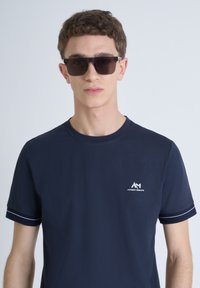 T-shirt di cotone blu navy con maniche corte, caratterizzato da cuciture bianche sui polsini e un piccolo logo sul petto. Indossato con occhiali da sole scuri.